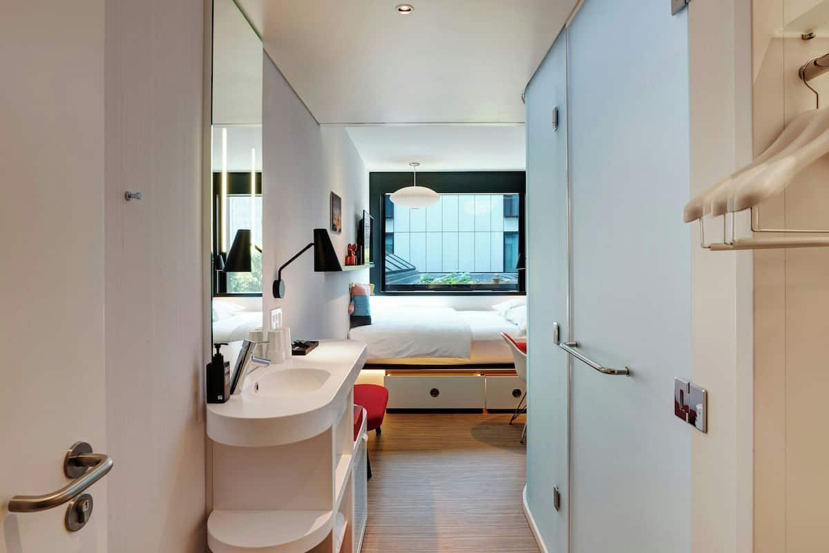A modern hotel room with a large bed, a bedside table, a mirror, a window, and a bathroom with a sink and toilet.