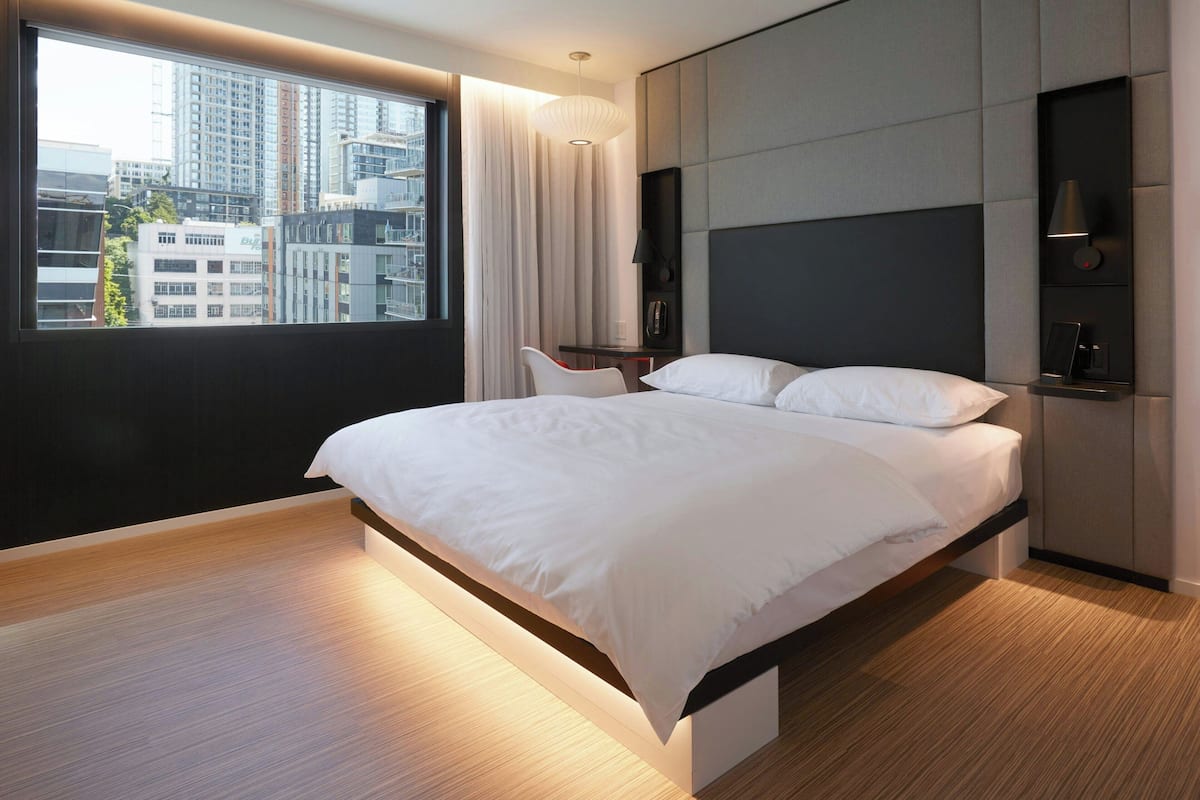 A modern hotel room with a large bed, a desk, and a view of the cityscape through the window.