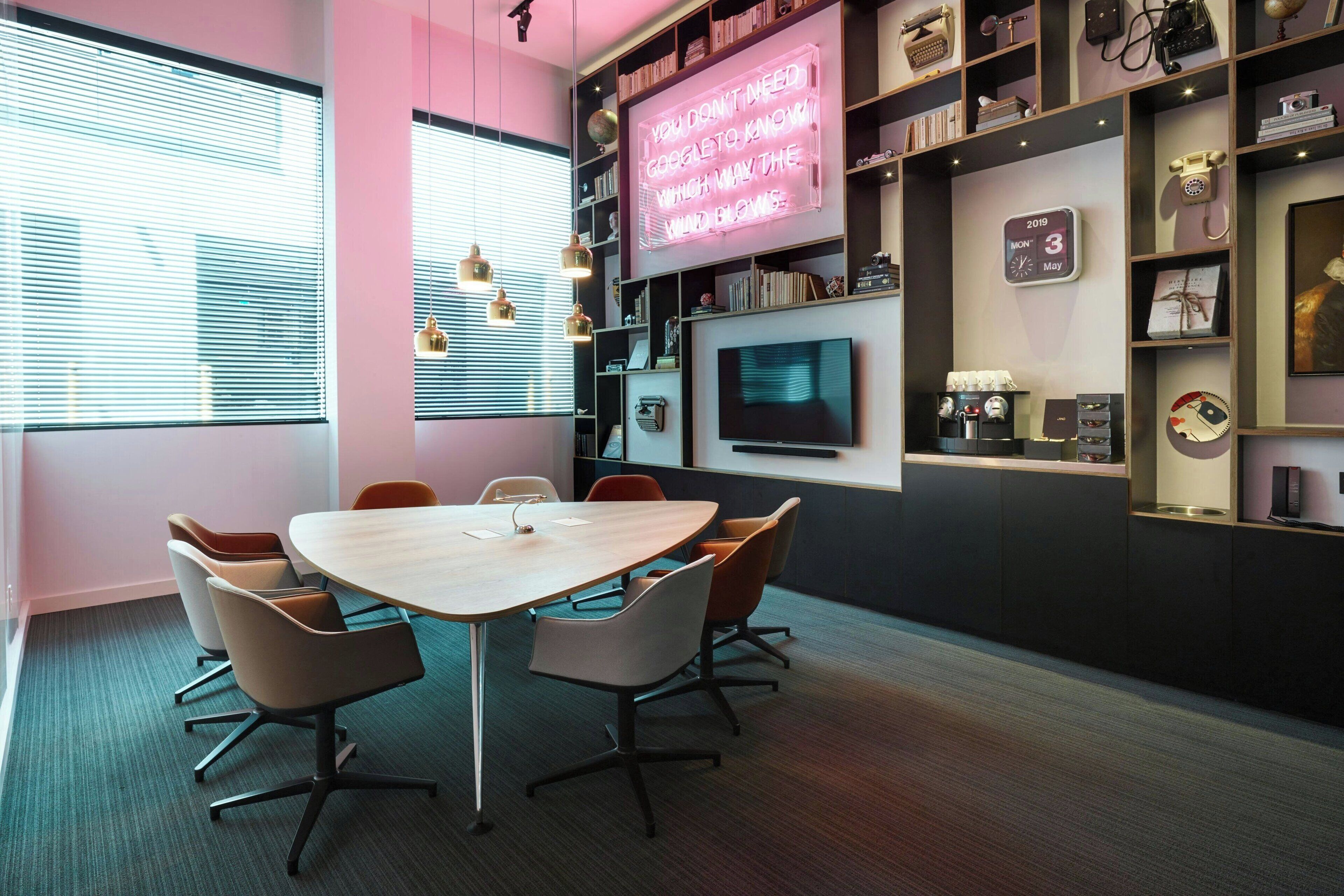A modern meeting room with a wooden table, comfortable chairs, a large TV, and shelves with various items.