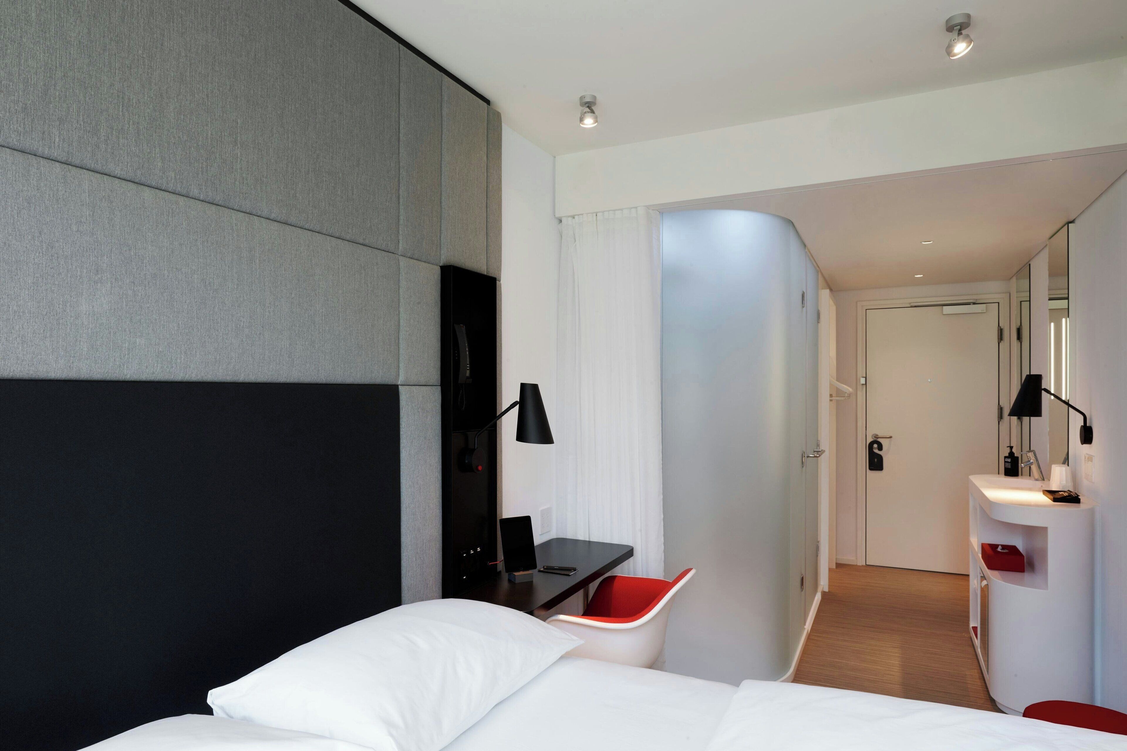 A modern hotel room with a bed, a desk, a red chair, a white sink, and a mirror.