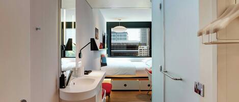 A modern hotel room with a large bed, a bedside table, a built-in desk, a window with a city view, and a bathroom with a sink and mirror.