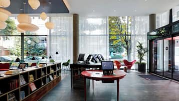 Una biblioteca moderna con estanterías, un ordenador portátil sobre una mesa y grandes ventanales con vistas a árboles y coches aparcados.