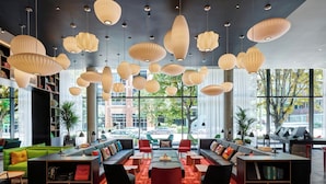 A modern lounge area with multiple seating options, including sofas and chairs, arranged around a central coffee table. There are bookshelves and a piano in the background. - citizenM Seattle South Lake Union (Seattle)