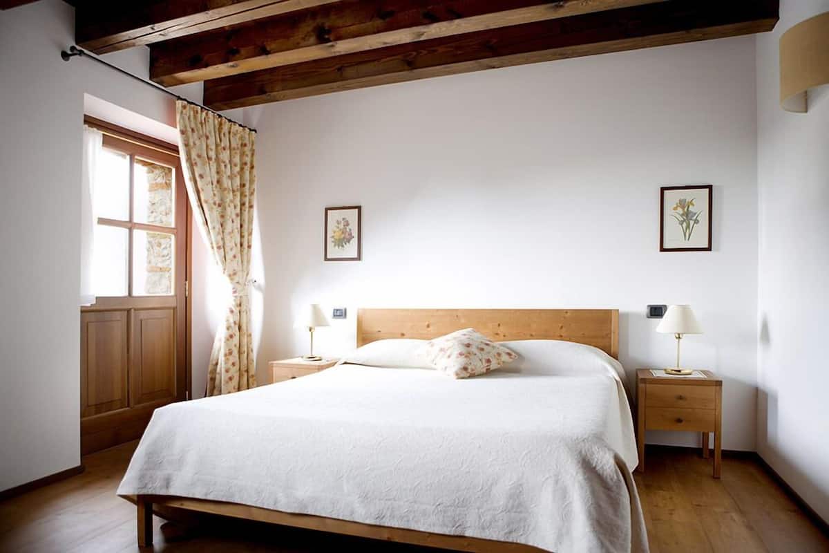 A bedroom with a wooden bed, two bedside tables with lamps, a window with curtains, and two framed pictures on the wall.