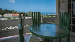 A balcony with a glass table and green chairs, overlooking a coastal view with houses and a clear sky.