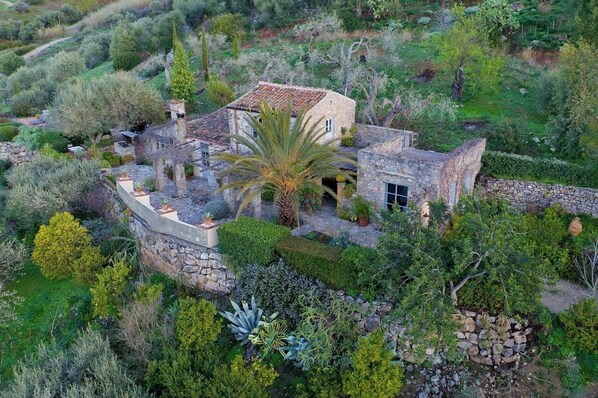 A stone house with a tiled roof surrounded by lush greenery and various plants.
