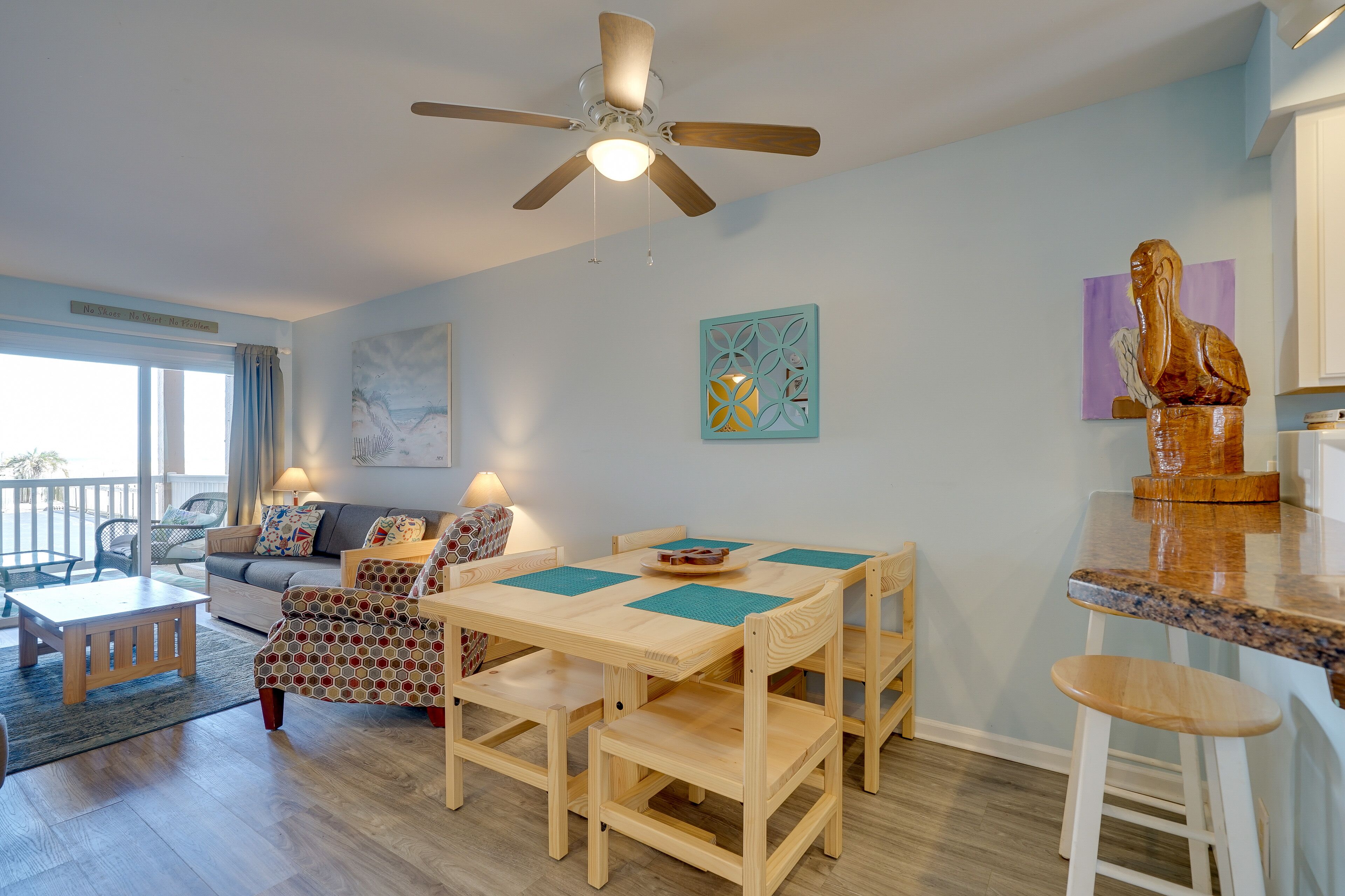 A spacious living area with a wooden dining table, chairs, and a ceiling fan. There is a sofa, a coffee table, and a decorative bird sculpture on the wall.
