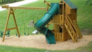 A wooden play structure with a slide and swings, surrounded by a grassy area.