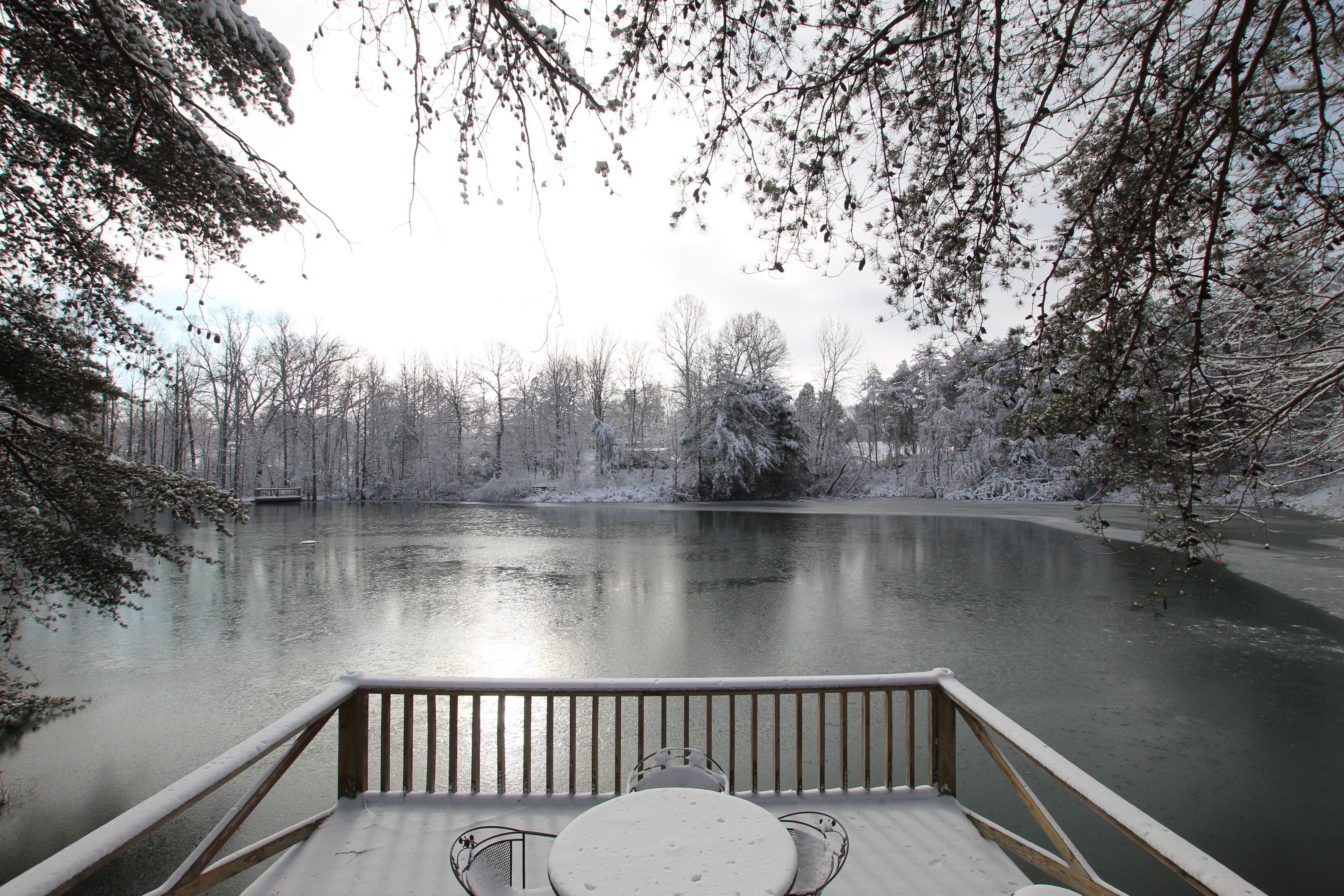 Un pontile di legno con tavolo e sedie che si affaccia su un lago ghiacciato, circondato da alberi innevati.