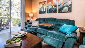 Ein Wohnzimmer mit einem türkisfarbenen Sofa, einem Couchtisch mit einer Ablage und einer Topfpflanze. An den Wänden hängen zwei Bilder und ein Fenster bietet Blick auf Bäume.