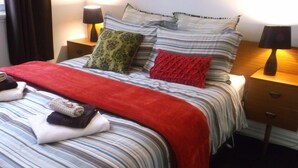 A neatly made bed with striped bedding, two pillows, and a red throw blanket. There are two bedside tables with lamps and a small drawer.