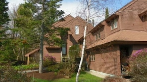 A two-story wooden house with a visible front porch, a small attached garage, and a well-maintained garden with various plants and trees. - Pet Friendly Ski Vacation Condo, Waterville Valley (Waterville Valley)