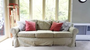 Un sofá color beige con tres almohadones, una ventana que da a vistas de árboles, y un reloj montado en la pared.