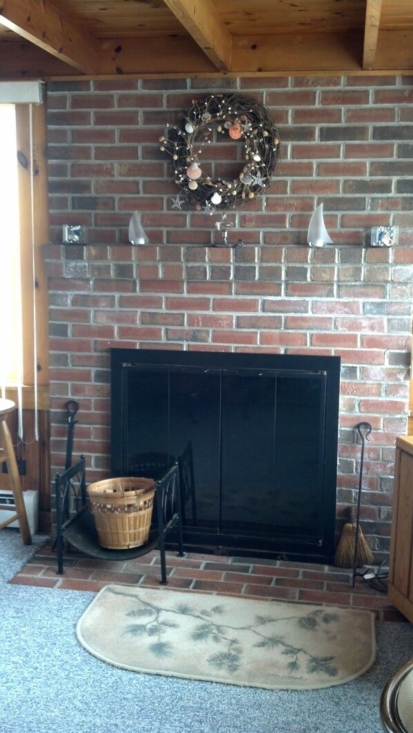 A brick fireplace with a decorative wreath above it, a black screen door, and a wooden chair and basket to the left.