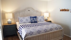 A neatly made bed with a patterned bedspread and pillows, flanked by two bedside tables with lamps, in a room with wooden flooring and a simple wall-mounted coat rack. - Tilghman Lakes By the Sea (North Myrtle Beach)