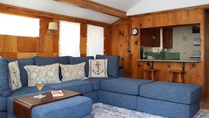 Un salon avec un canapé d’angle, un plafond en bois, un coin cuisine doté d’un plan de travail vert et une horloge au mur.
