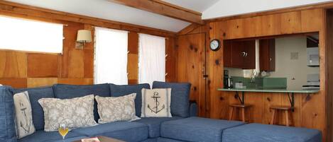 Ein Wohnzimmer mit Ecksofa, Holzboden und einer Küchenzeile mit grüner Arbeitsplatte. An der Wand hängt eine Uhr.