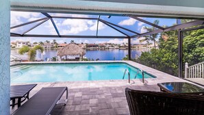 Une piscine avec vue sur un lac et des maisons, un patio couvert avec des chaises et une structure à toit de chaume.