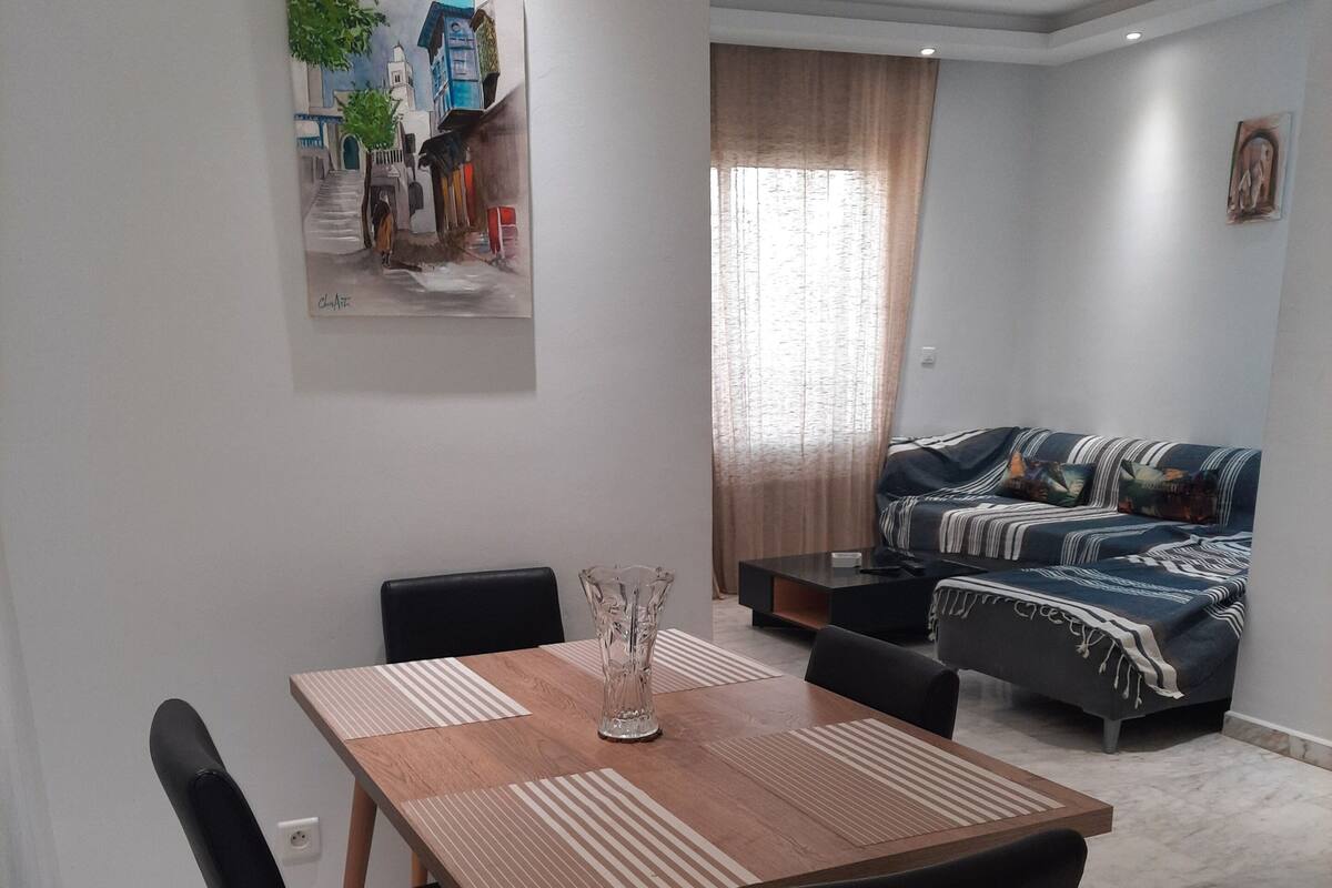 Un comedor con mesa de madera y sillas negras, una sala de estar con sofá y cortinas, y un cuadro en la pared.