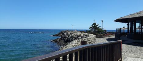 A coastal walkway with a wooden railing, a rocky outcrop, and a thatched-roof structure.