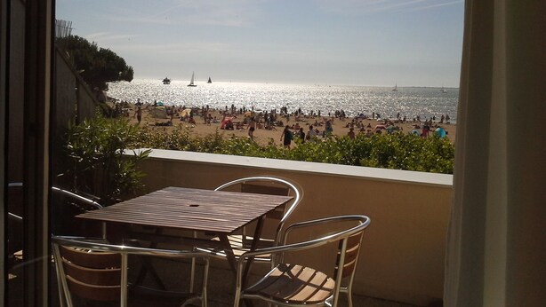 Un balcón con mesa y sillas que da a una playa concurrida y varios veleros.