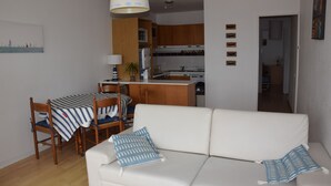 Una sala de estar moderna con un sofá blanco y cojines a rayas, un comedor con mantel a rayas, sillas de madera y una cocina con armarios de madera y un microondas.