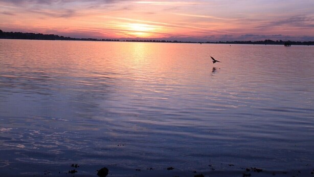 Ein ruhiger See spiegelt einen Sonnenuntergang wider, der bewölktem Himmel entgegenblickt.