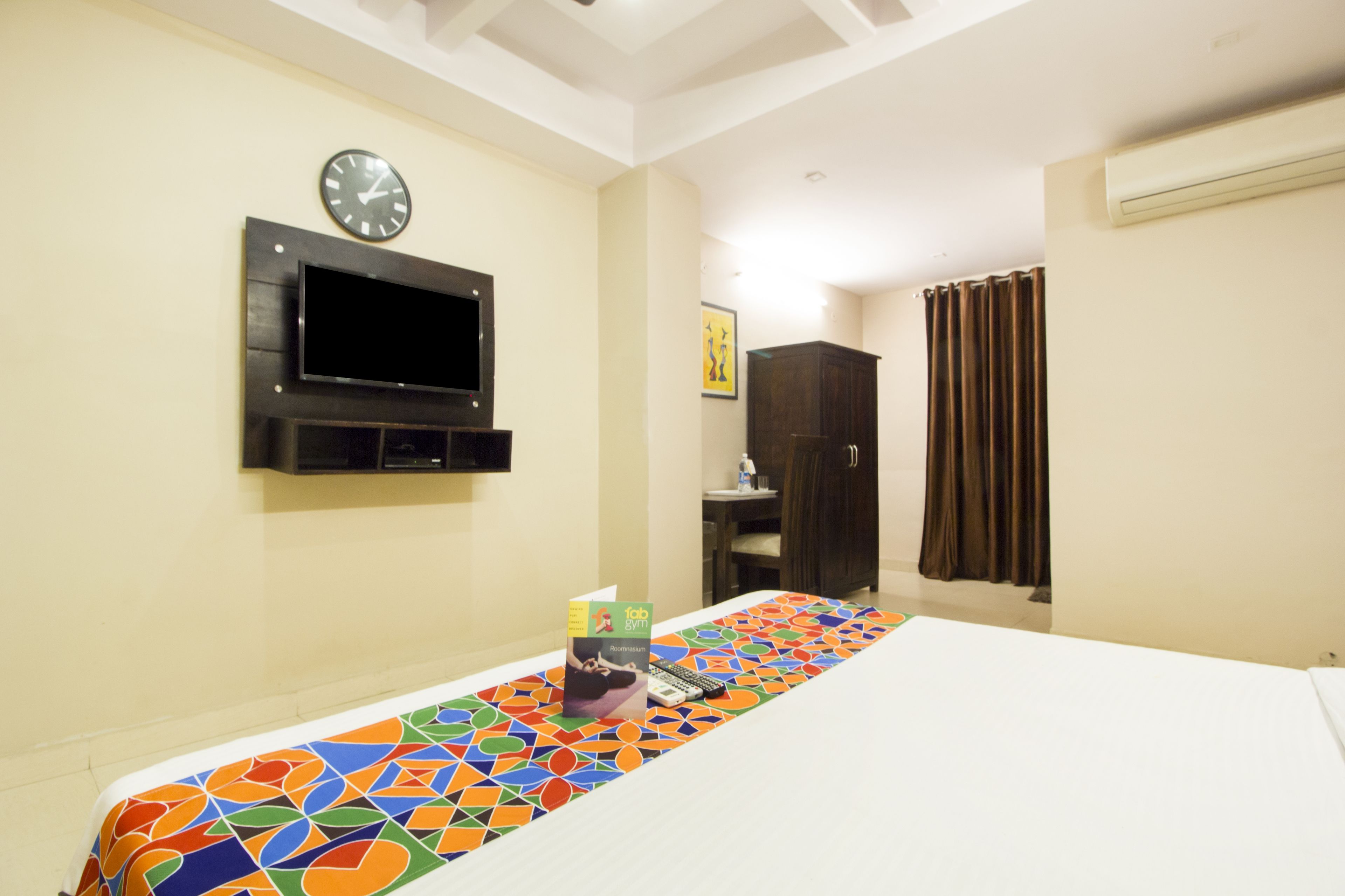 Habitación de hotel con cama, televisor montado en la pared, un reloj y ventana con cortinas.