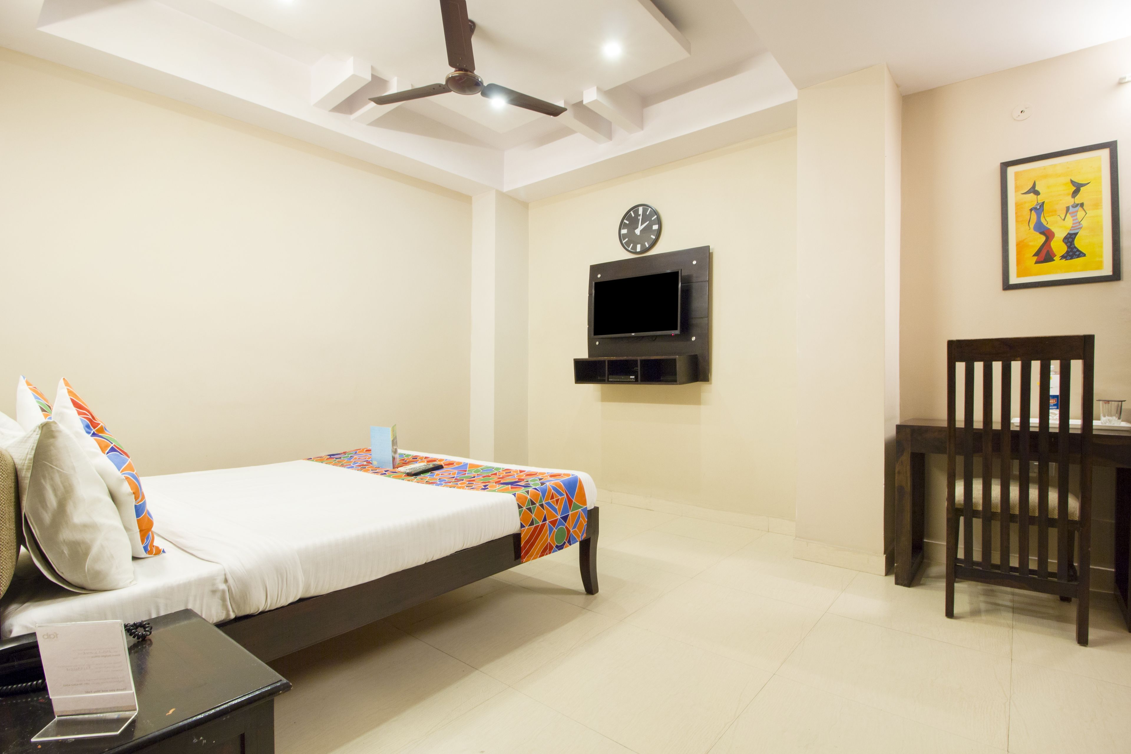 A hotel room with a bed, a desk, a chair, a TV mounted on the wall, and a ceiling fan.