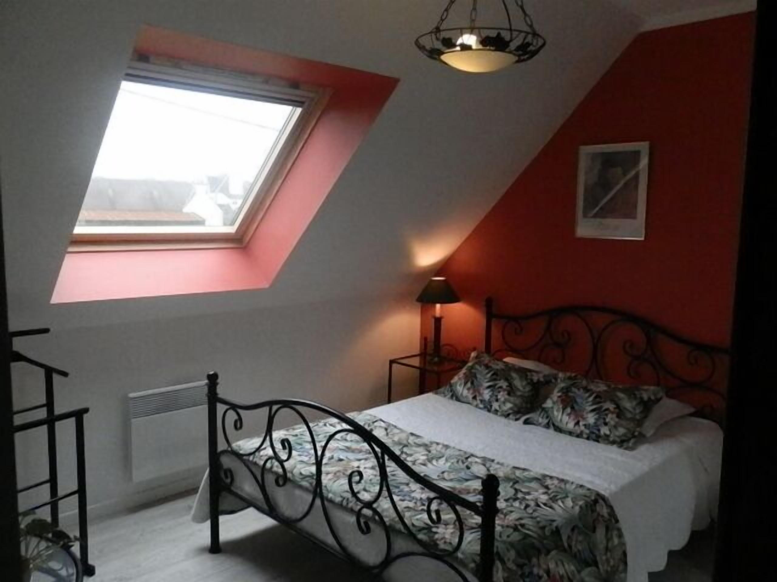 Une chambre avec un plafond en pente, un puits de lumière, un lit recouvert d’une couvre-lit à motifs floraux, une lampe de chevet et un radiateur.