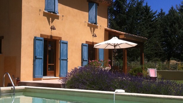 A pool area with a clear pool, a building with blue shutters, and a patio with an umbrella.
