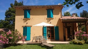 A two-story building with a tiled roof, a covered patio with a table and chairs, and a wooden lounge chair.