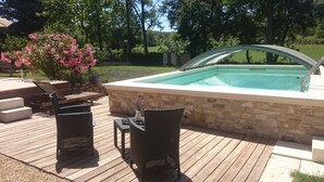 A swimming pool with a stone wall and a wooden deck. There are two wicker chairs and a table on the deck.