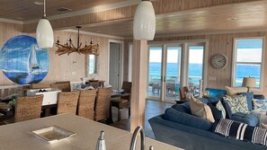 Amplia sala de estar con un gran ventanal con vista al océano, comedor con mobiliario de mimbre y cocina con una isla grande.