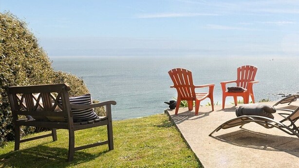 Eine Terrasse mit einer Holzbank, zwei roten Adirondack-Stühlen und einem Liegestuhl, der auf ein Gewässer ausgerichtet ist.