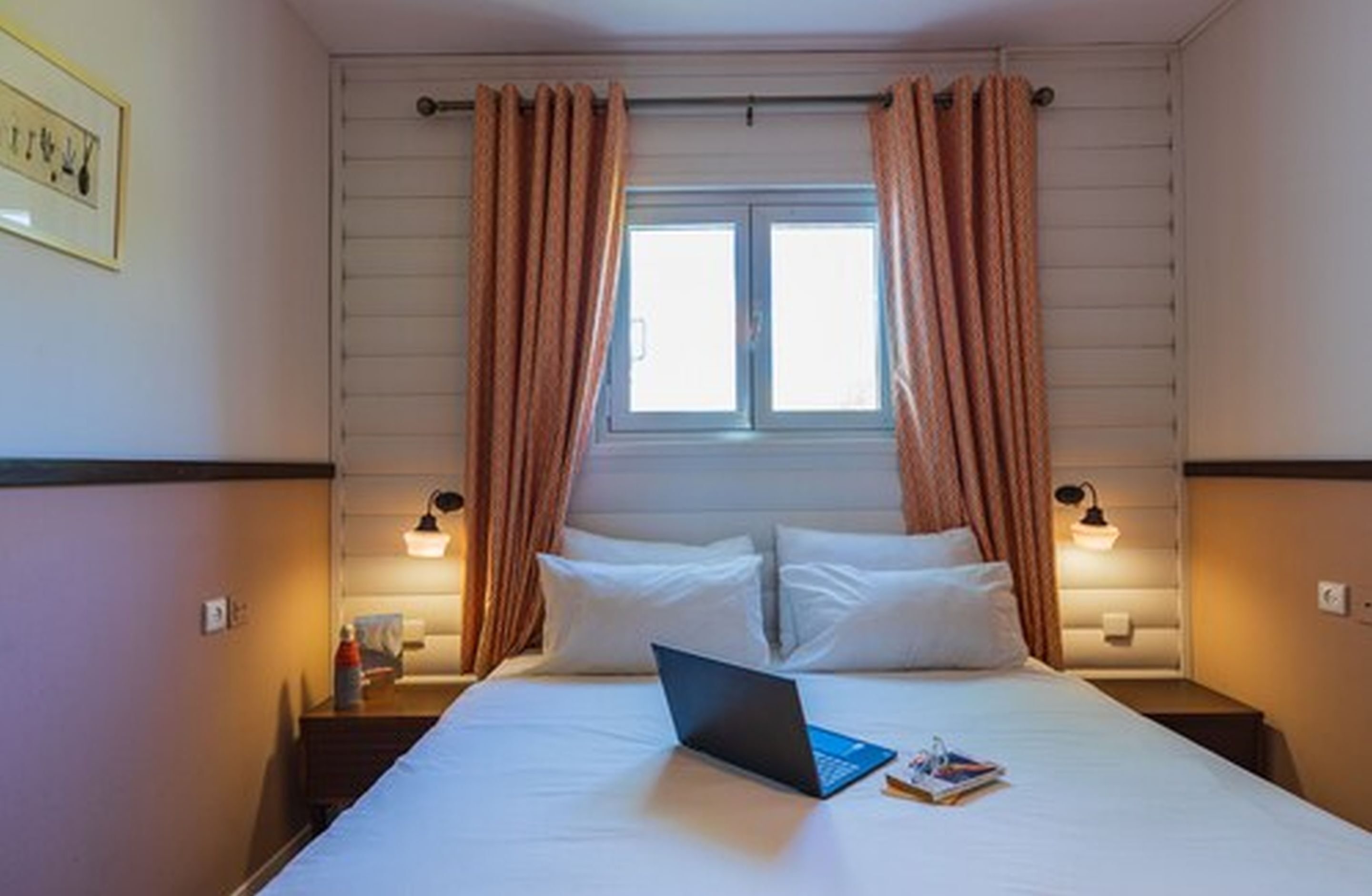Una habitación de hotel con una cama, una computadora portátil y un ventanal con cortinas.