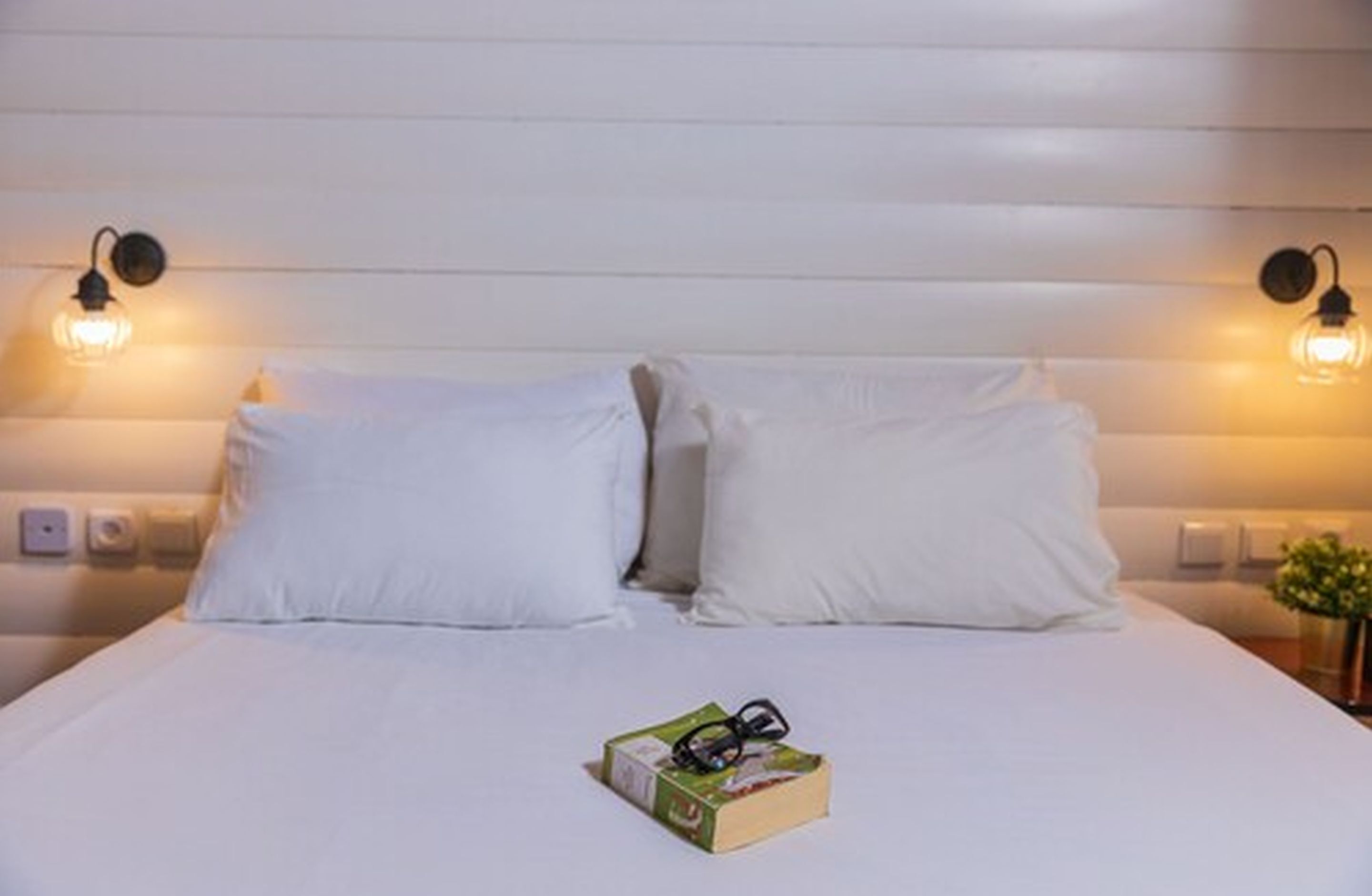 A neatly made bed with white linens and pillows, a small bedside table with a book and glasses, and two wall-mounted light fixtures.