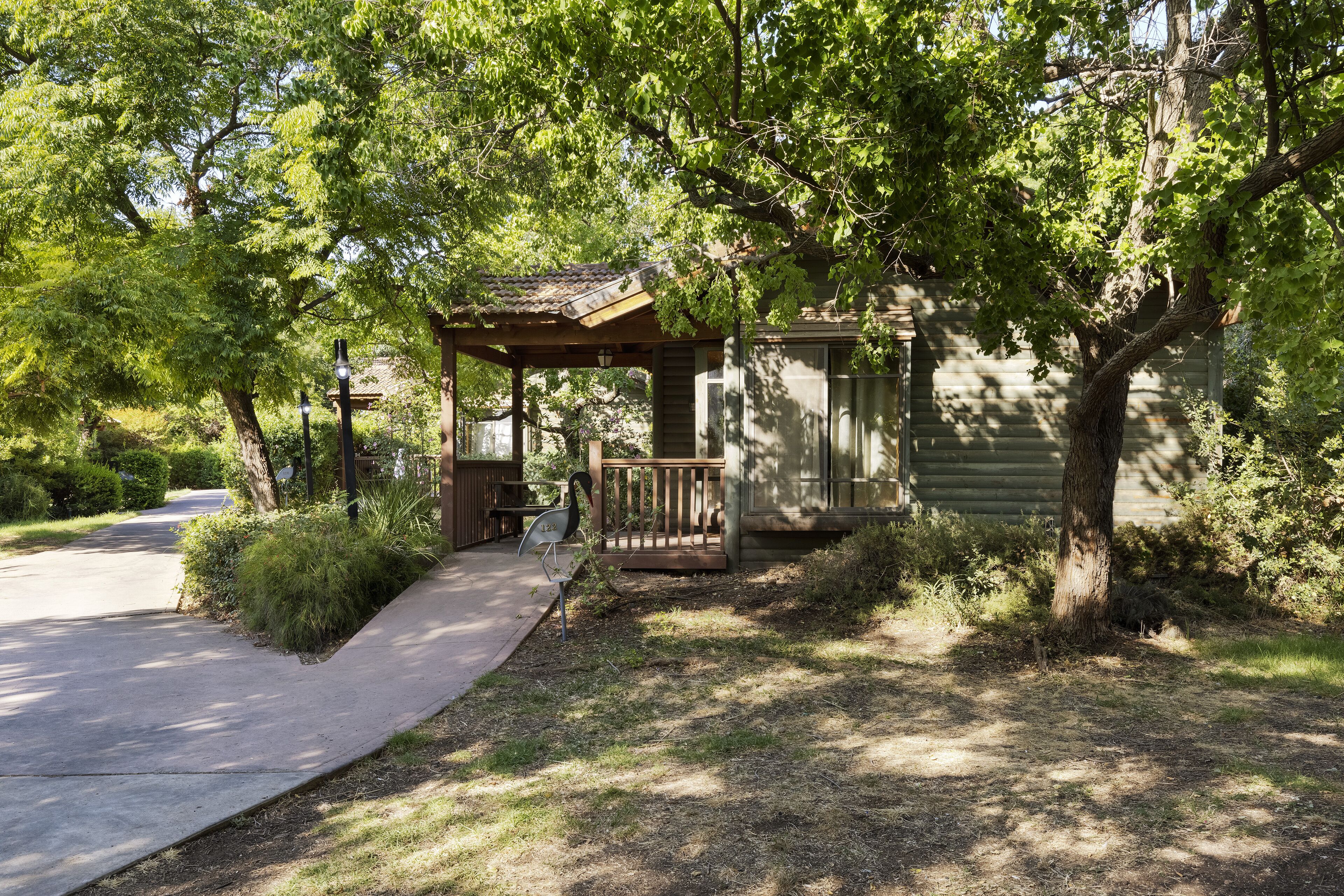 A covered outdoor seating area with a wooden bench and a small table, surrounded by trees and a building with a sloped roof.