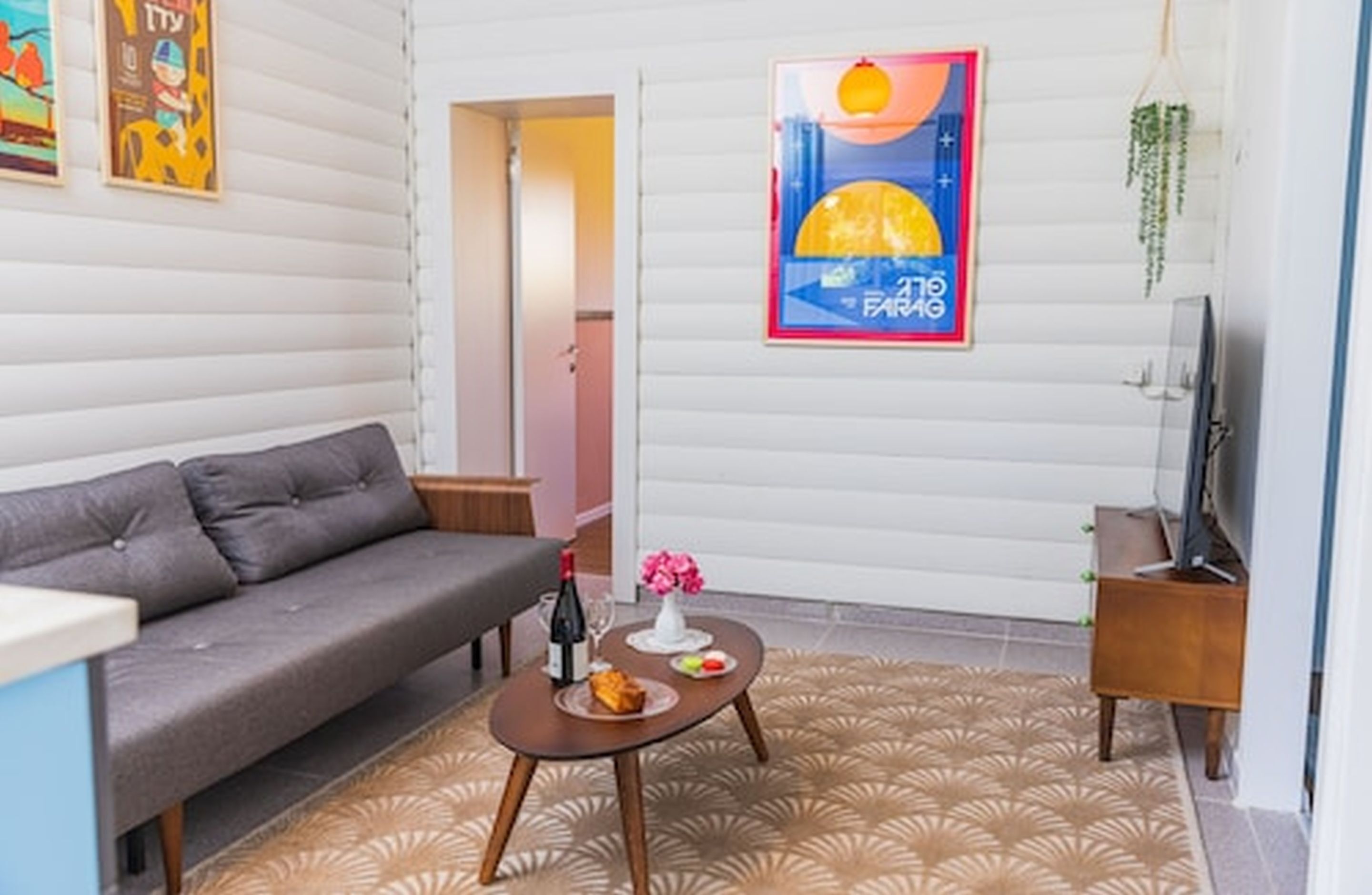 Ein modernes Wohnzimmer mit einem grauen Sofa, einem kleinen Couchtisch mit einer Weinflasche und Blumen sowie einem Fernseher auf einem Holzgestell.
