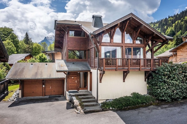 A two-story wooden chalet with a balcony, a garage, and a mountain backdrop.
