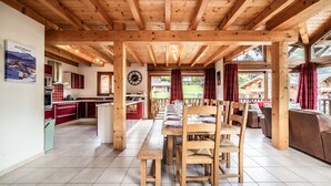 Eine geräumige Küche mit Holzbalken, einem Essbereich mit langem Tisch und Stühlen und Blick auf einen Garten durch große Fenster.