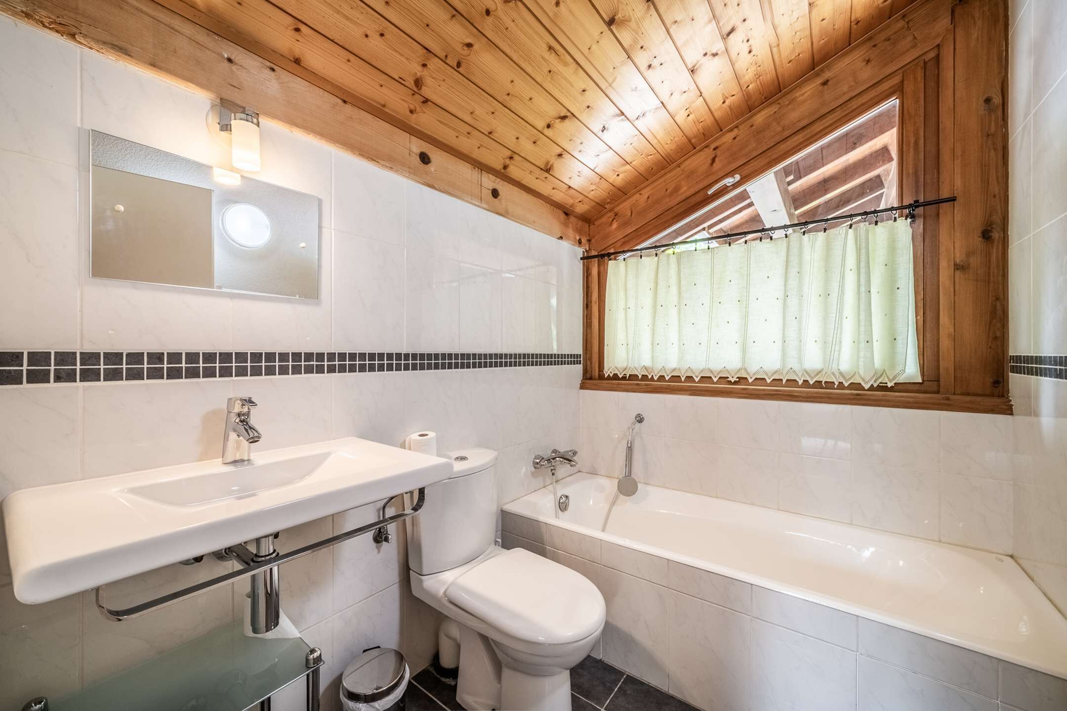 A bathroom with a wooden ceiling, white tiles, a sink, a toilet, and a bathtub.
