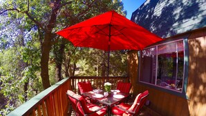 Une terrasse en bois avec une table et des chaises, un parasol rouge et une fenêtre donnant sur une zone boisée.