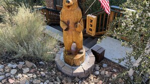 Une statue d’ours en bois tenant un drapeau américain, entourée de rochers et de plantes, avec une terrasse en bois et une maison en arrière-plan.