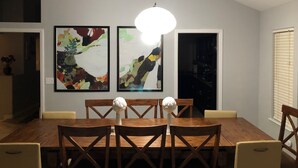 Een eetgedeelte met een houten tafel en stoelen, twee abstracte schilderijen aan de muur, en een hanglamp.