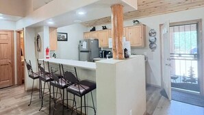 Une cuisine moderne avec des poutres en bois au plafond, un îlot central doté de tabourets de bar et une vue sur l’extérieur grâce à une porte vitrée.