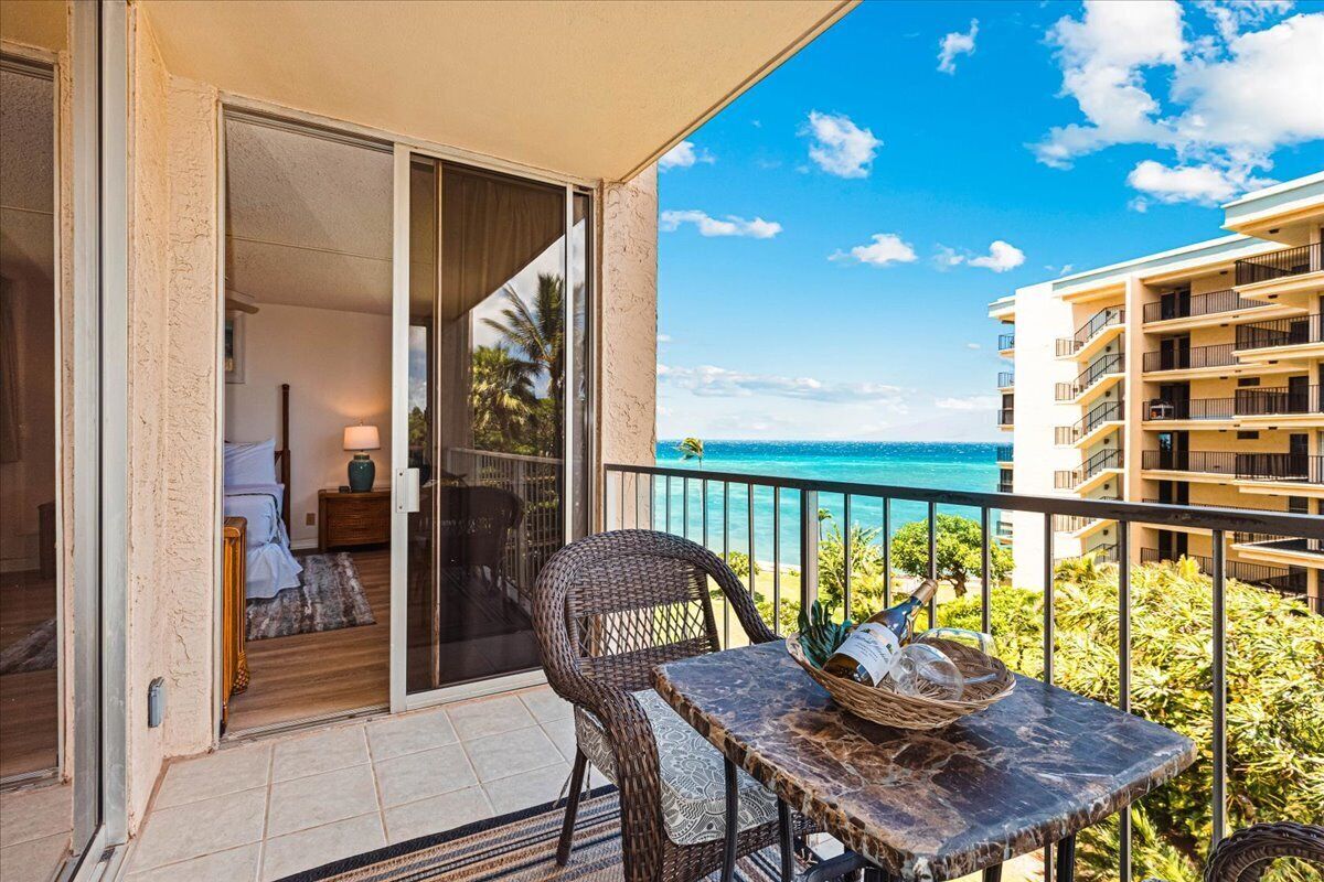 Een balkon met een tafel en rieten stoel, met uitzicht op een strand en een gebouw met meerdere balkons.