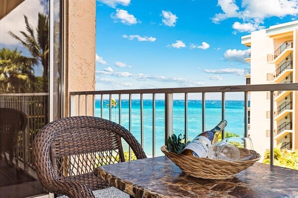 A wicker chair and table with a bottle and glasses on a balcony overlooking the ocean.