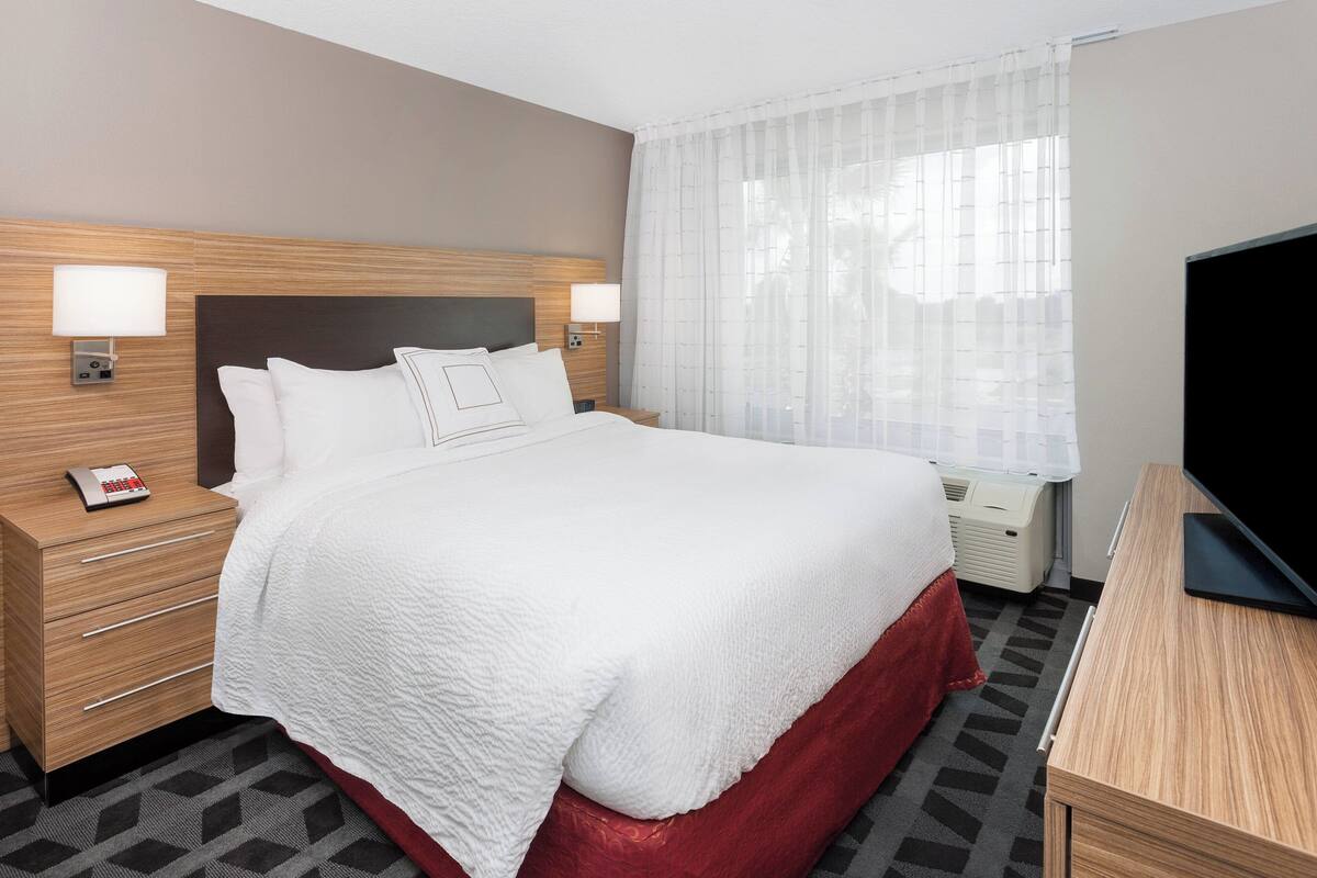 A hotel room with a large bed, two bedside tables with lamps, a TV, and a window with sheer curtains.