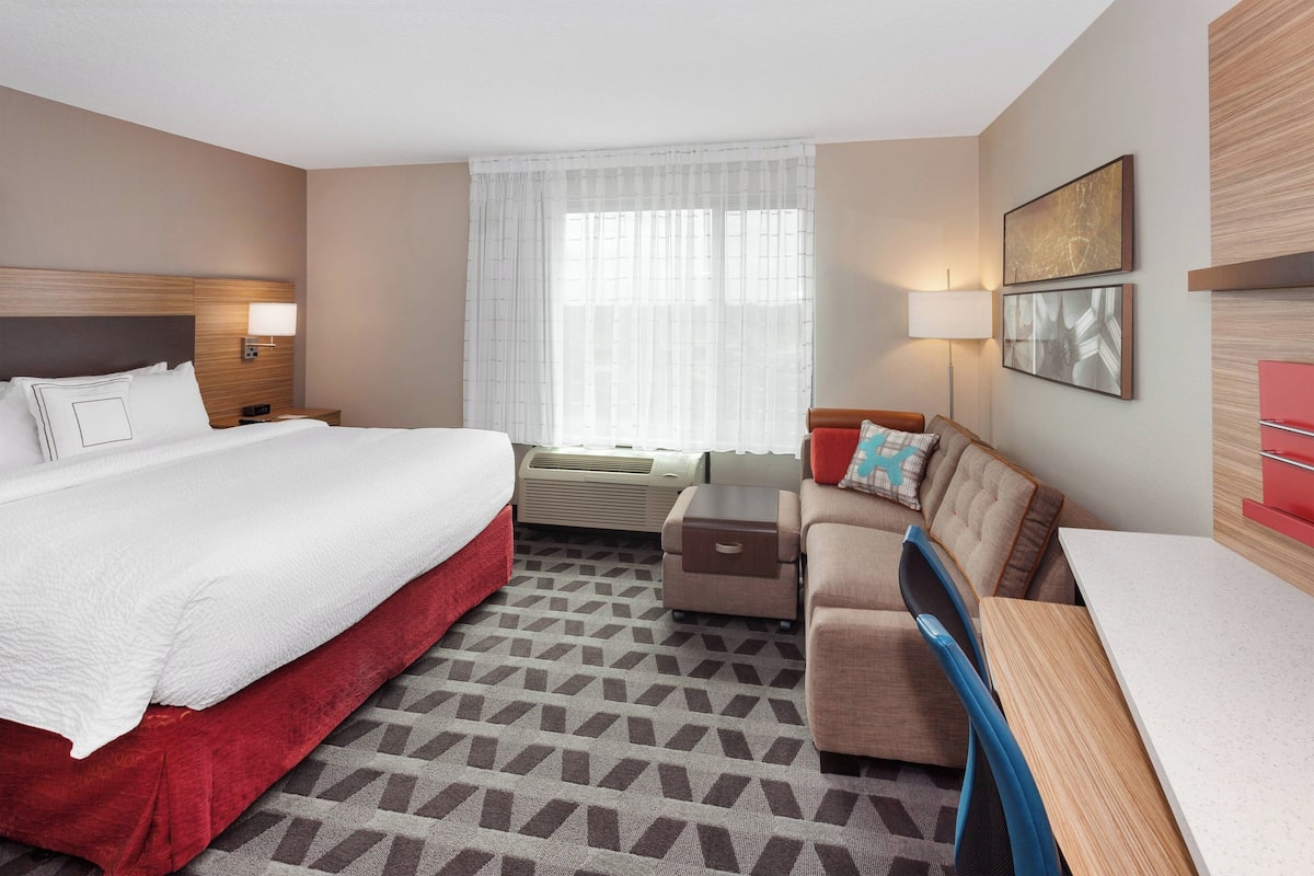 A hotel room with a large bed, a sofa, a small table, and a desk. There is a window with curtains, a wall-mounted lamp, and two framed pictures.